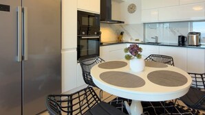 Een kleine keuken met een koelkast, oven en magnetron. Er staat een eettafel met stoelen en een kleine potplant.