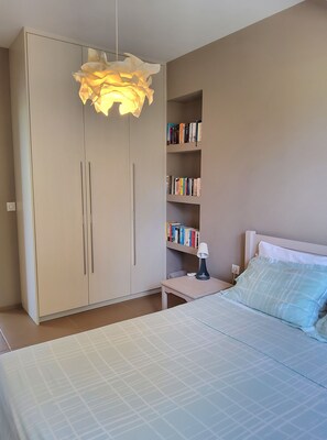 Un dormitorio con cama, mesita de noche con lámpara, armario y estantería.