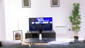 Ein modernes Wohnzimmer mit Flachbildfernseher, gerahmten Bildern an der Wand, einer Topfpflanze und einer Fernbedienung auf einem Couchtisch.