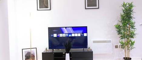 A modern living room with a flat-screen TV, framed pictures on the wall, a potted plant, and a remote control on a coffee table.