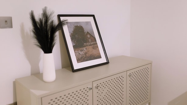 Ein gerahmtes Foto auf einer Kommode, eine weiße Vase mit dunkelgrünen Blättern und ein Lichtschalter.
