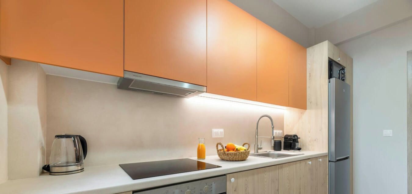 A modern kitchen with orange cabinets, a stainless steel refrigerator, a coffee maker, and a fruit basket.