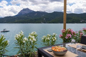 A dining table with a bowl of food, a wicker chair, and a view of a lake with mountains in the background. - Mobile Homes for 4 People With Lake View in Residence L'ecrin du Lac, Chorges (Chorges)