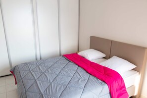Ein Bett mit grauer Bettdecke und einer pinkfarbenen Zudecke, weißen Kissen und einem hölzernen Kopfteil in einem Zimmer mit weißen Wänden und einem gefliesten Boden.