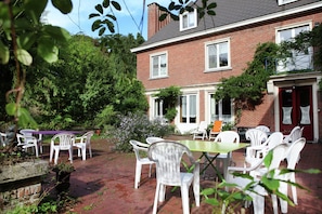 A brick building with a patio area featuring white plastic chairs and tables, surrounded by greenery. - Spacious 11-bed Family Retreat (Ruiselede)