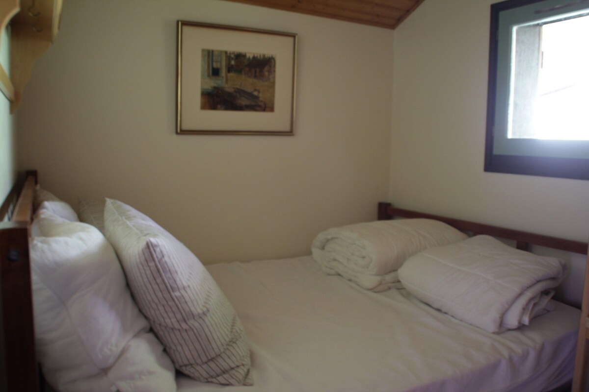 A single bed with pillows and a folded blanket, a framed picture on the wall, and a window with a view of the outside.