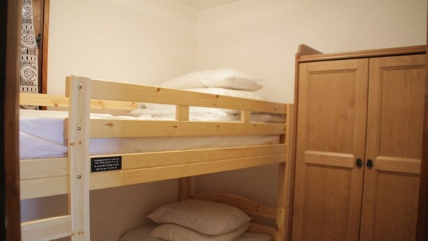 Uma cama beliche com cabeceira de madeira, a cama inferior com almofadas e um guarda-roupa de duas portas.