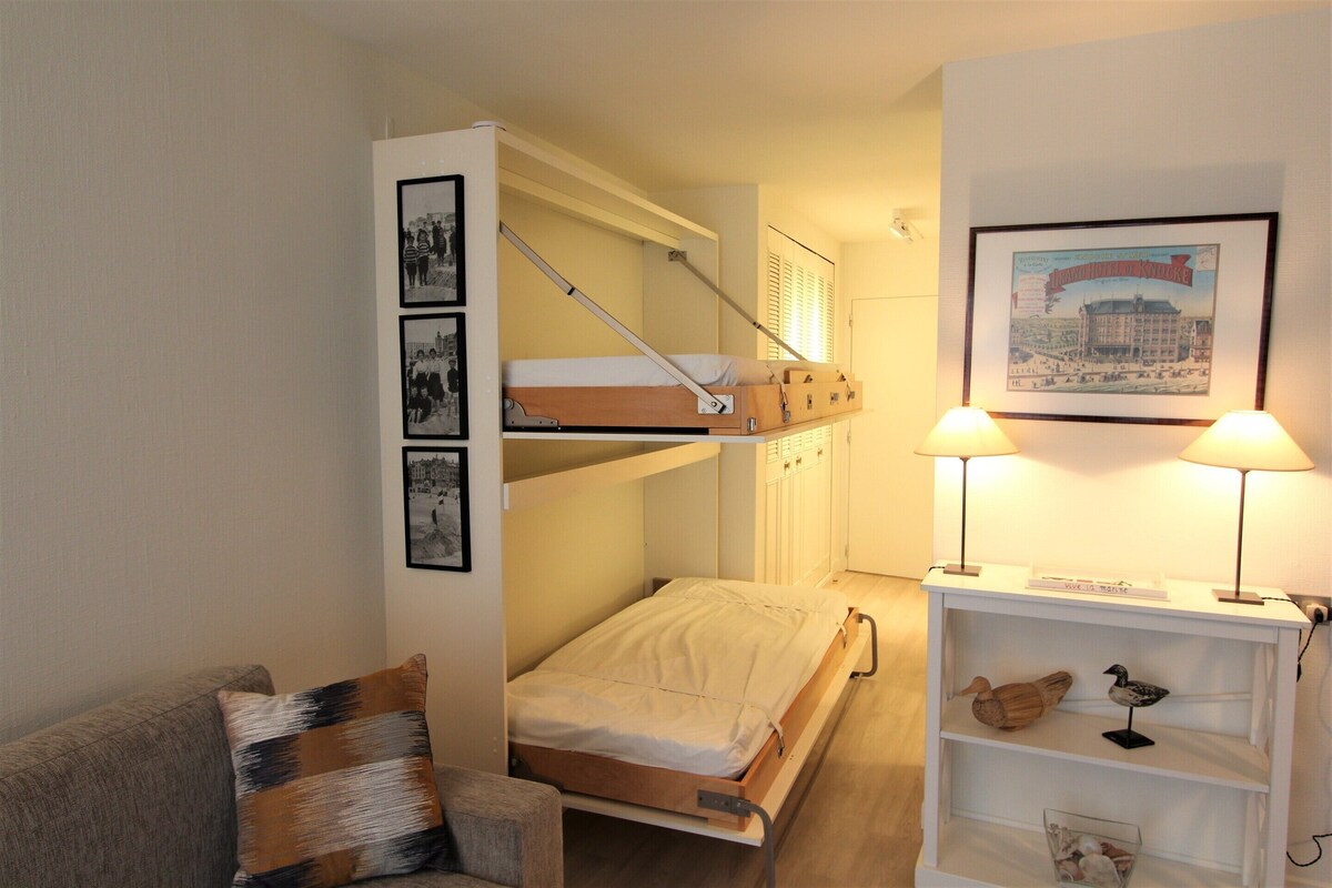Una habitación de hotel compacta con una cama litera, un pequeño sofá, una mesita de noche con lámpara y una estantería con objetos decorativos.