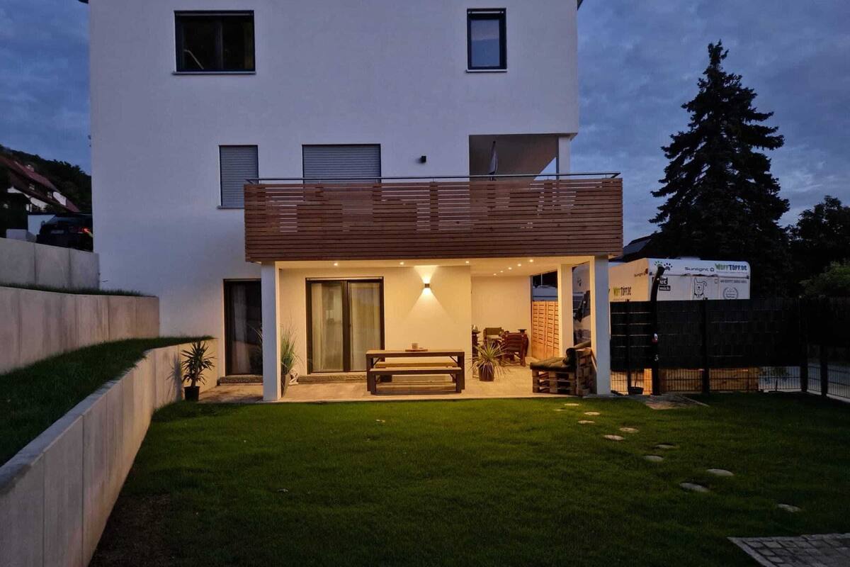 Ein modernes, zweistöckiges Haus mit Balkon, einer Terrasse mit Tisch und Stühlen und einem gepflegten Rasen.