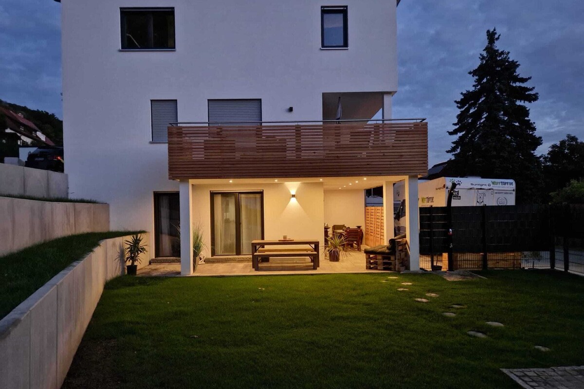 Et moderne hus med to etager, en balkon, en patio med bord og stole, samt en velplejet græsplæne.
