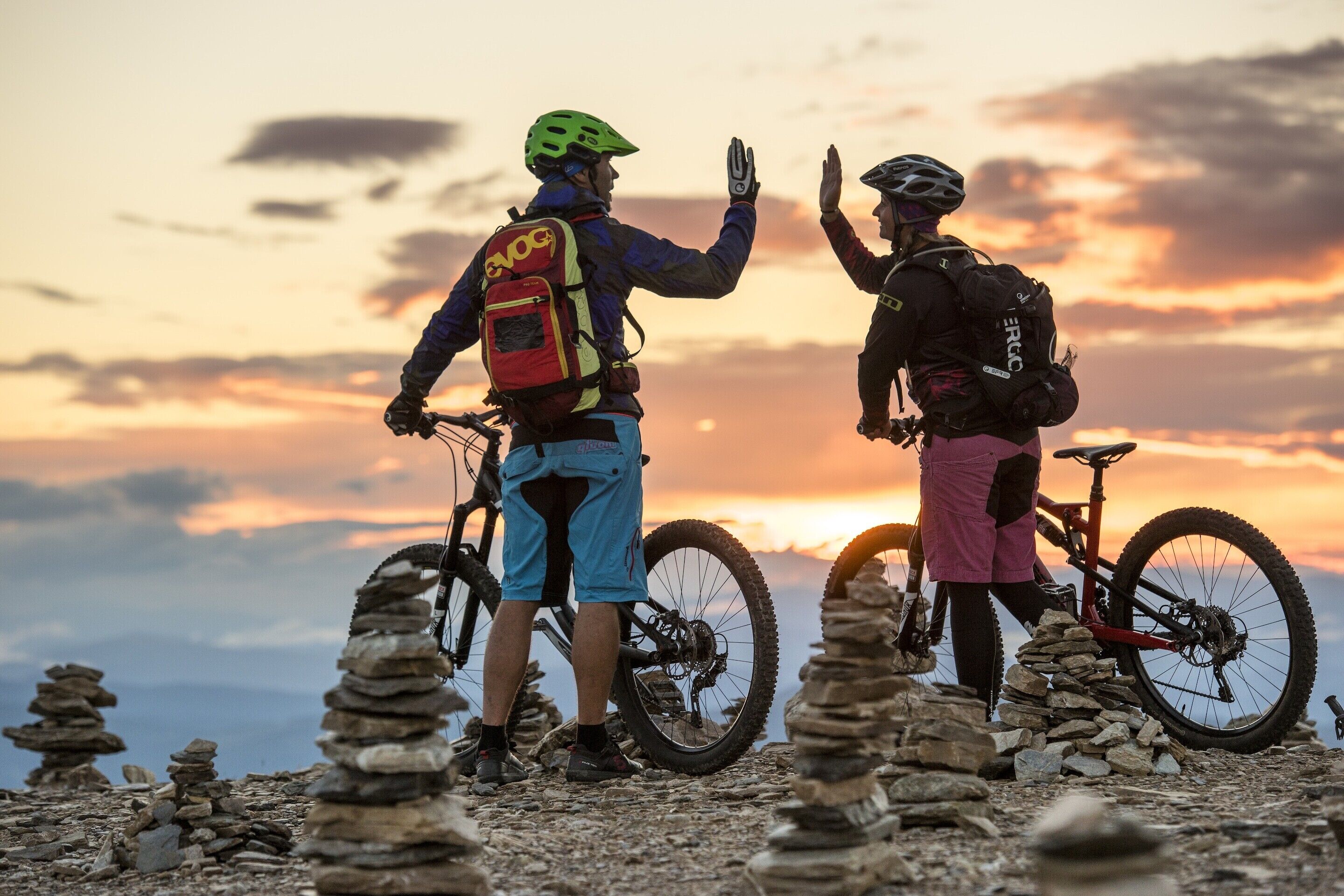 Dos personas con bicicletas, un atardecer y formaciones rocosas apiladas.