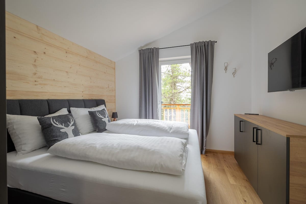 Un dormitorio moderno con cabecera de madera, una cama grande con ropa blanca, cortinas grises, un televisor montado en la pared y un mueble de madera.
