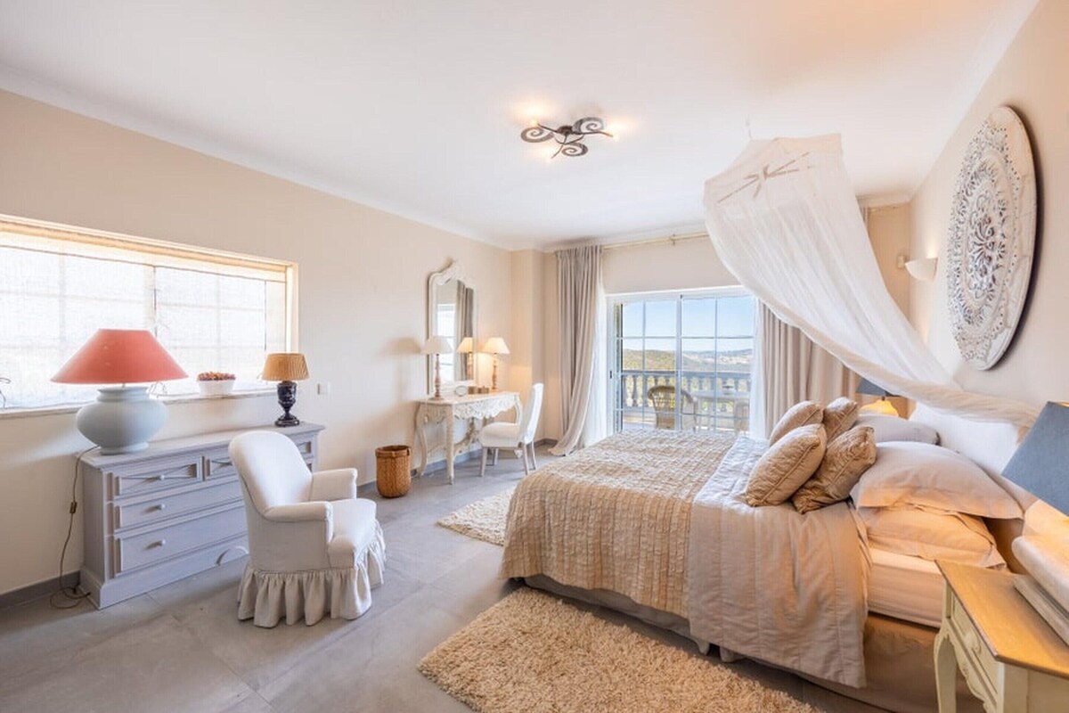 A bedroom with a bed, a desk, a chair, a dresser, and a large window with curtains.