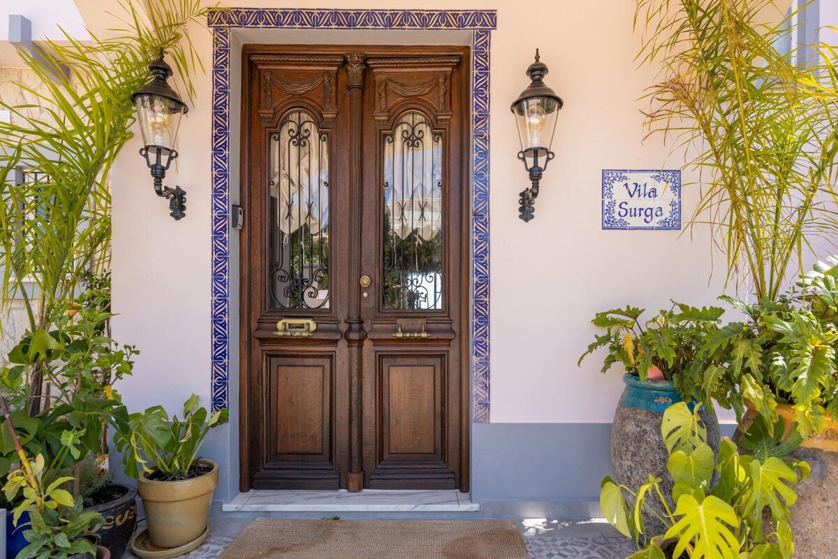 A wooden double door with glass panels, flanked by two wall-mounted lamps, and a sign reading 'Villa Surga' on the wall.