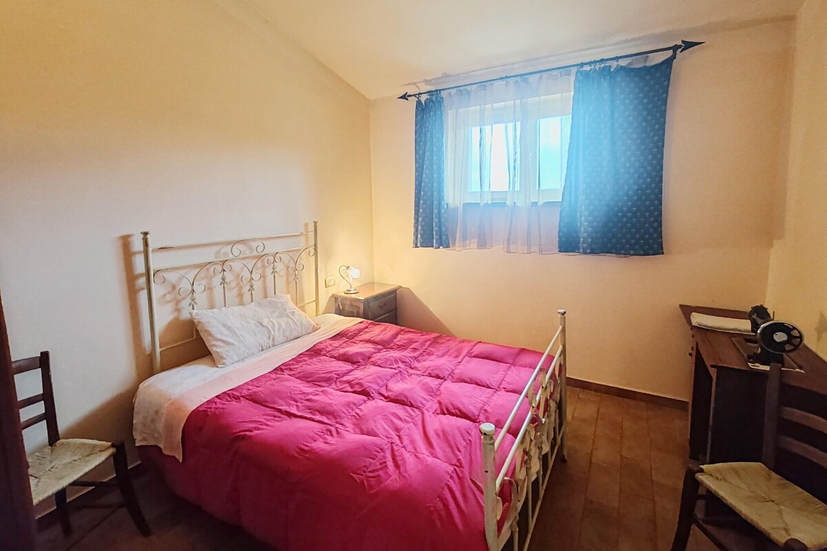 Ein Schlafzimmer mit einem Bett, einem Sessel, einem Schreibtisch und einem Fenster mit Vorhängen.