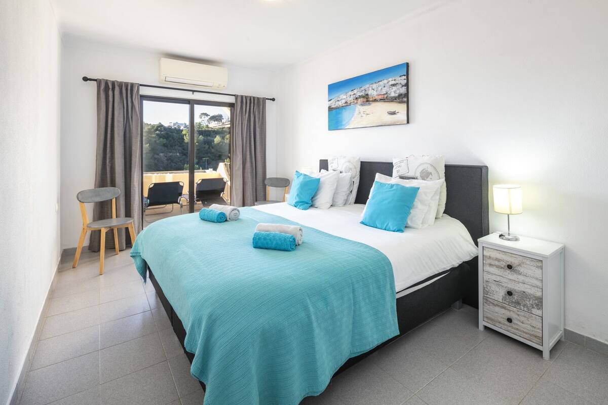 A bedroom with a bed, a nightstand, a chair, a window with curtains, and a painting on the wall.