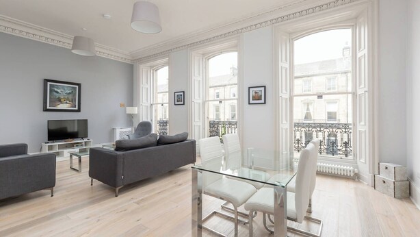A modern living room with a grey sofa, a glass dining table with white chairs, a flat-screen TV, and large windows with a view of buildings. - 6 on Chester Apartments (Edinburgh)