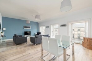 Una sala de estar moderna con un televisor de pantalla plana, sofás grises, una mesa de comedor de vidrio y ventanas amplias que ofrecen vistas a edificios.