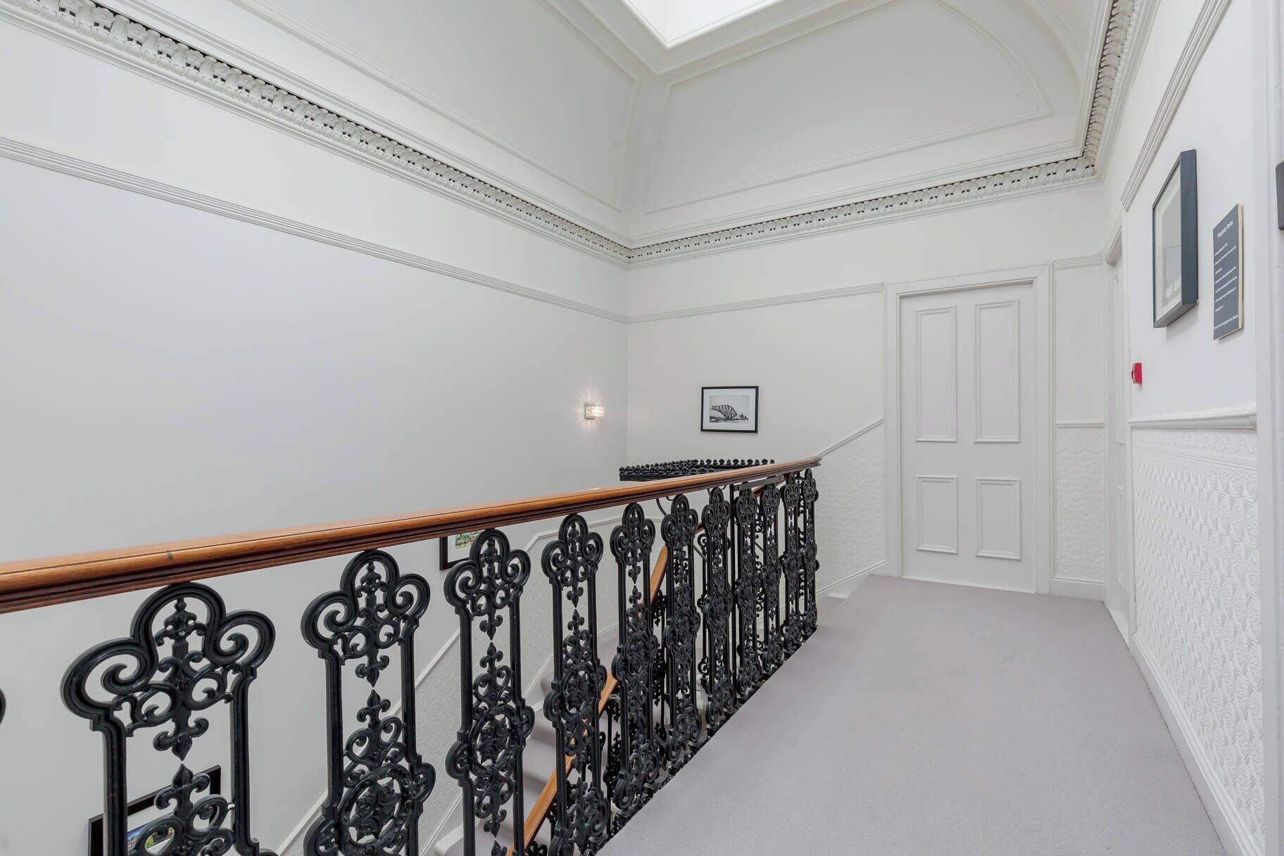 Un intérieur blanc avec une rampe d’escalier en bois, un tableau encadré au mur et une porte.