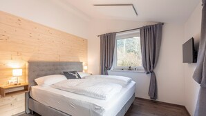 A bedroom with a bed, bedside table, lamp, window with curtains, and a flat-screen TV. - Chalet in St. Englmar With Private Sauna & Hottub (Sankt Englmar)