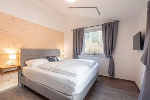 Ein Schlafzimmer mit Bett, Nachttisch, Lampe, Fenster mit Vorhängen und einem Flachbildfernseher.