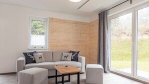 A modern living room with a sectional sofa, two ottomans, a wooden coffee table, and large windows offering a view of the outdoors. - Chalet in St. Englmar With Private Sauna & Hottub (Sankt Englmar)