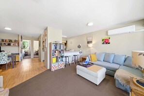 Ein Wohnzimmer mit einer Couch, einem Couchtisch, einem Bücherregal und einer Küche im Hintergrund.