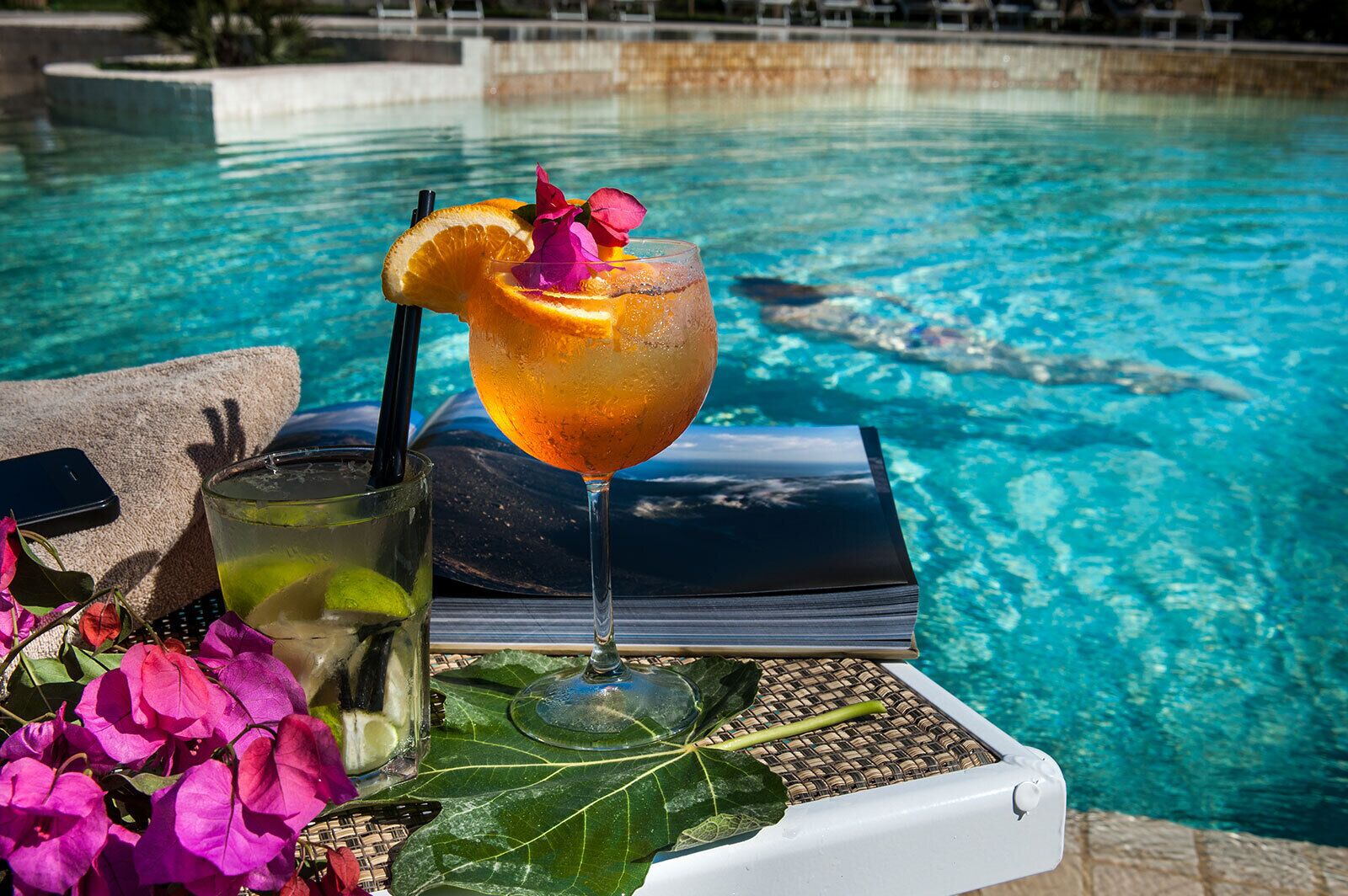 Un vaso de bebida con una rodaja de naranja y una flor, un vaso con pajita y un libro sobre una bandeja junto a una piscina.