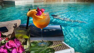 Un vaso de bebida con una rodaja de naranja y una flor, un vaso con pajita y un libro sobre una bandeja junto a una piscina.