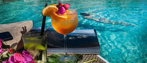A glass of drink with a slice of orange and a flower, a tumbler with a straw, and a book on a tray by a pool.