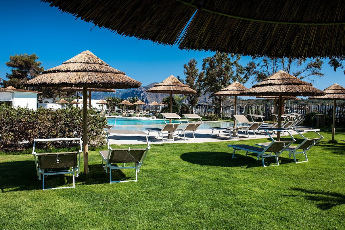 Un área junto a la piscina con sillones de descanso y sombrillas de paja, rodeada de follaje y un cielo despejado.