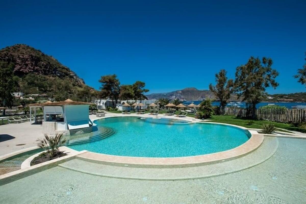 Una piscina de diseño circular, rodeada de sillones de descanso y sombrillas, con vistas a colinas y una masa de agua al fondo.