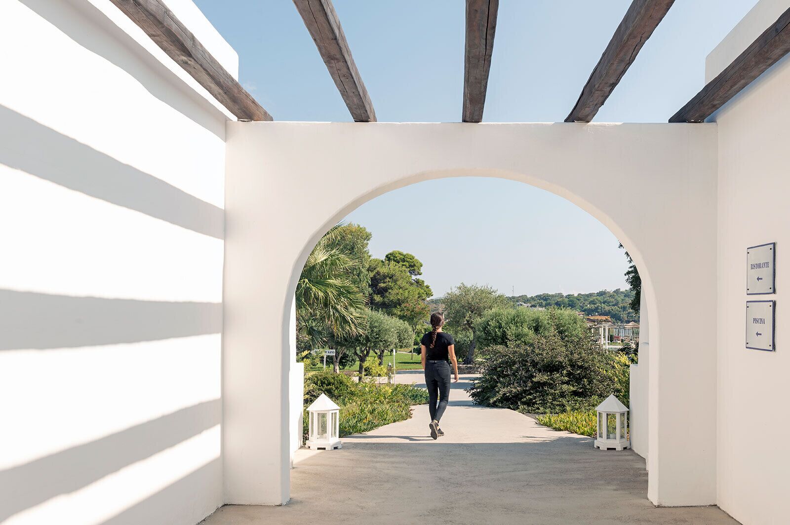A person walking through a white archway with a view of greenery and buildings in the distance.