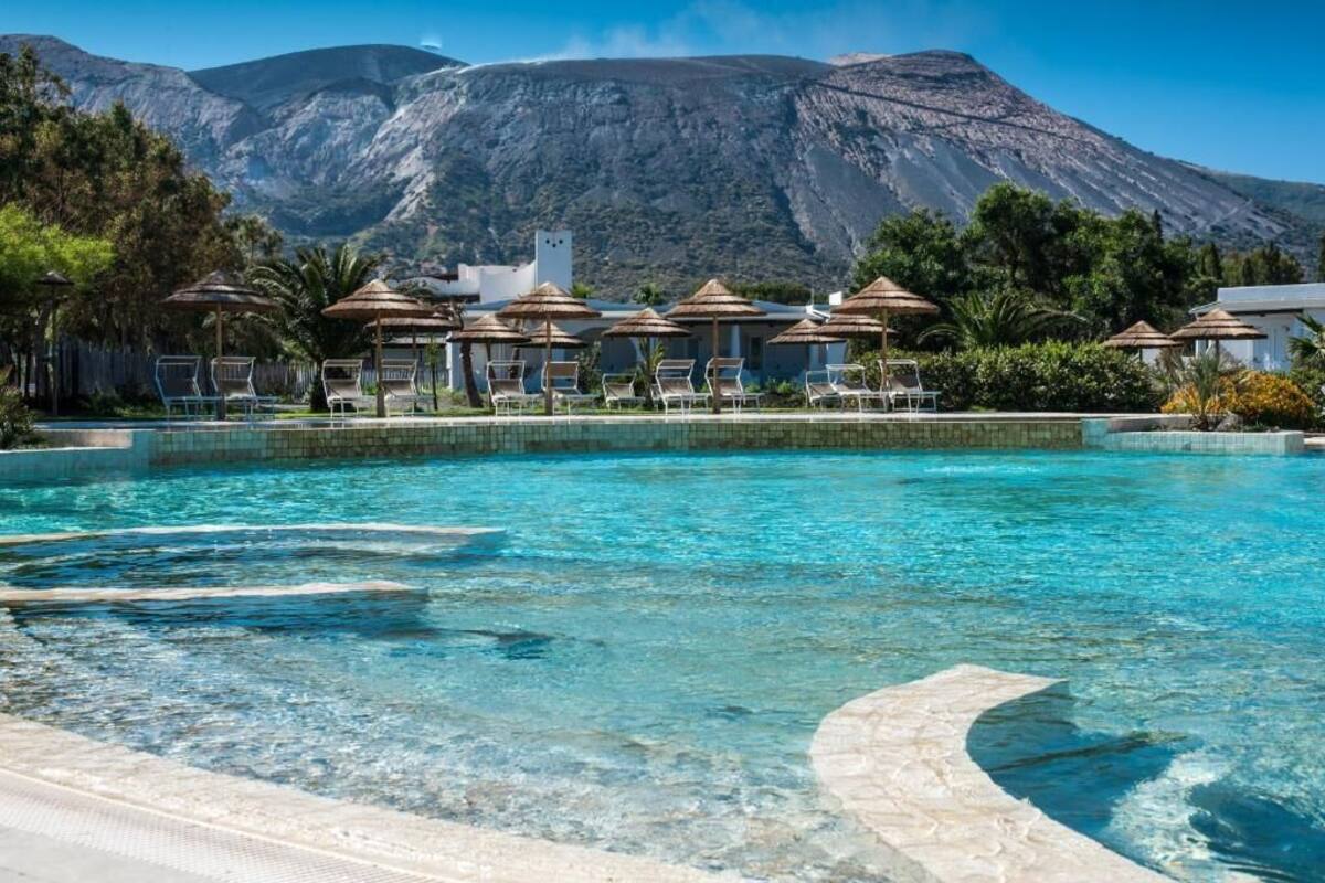 Una piscina con agua cristalina, sillas de descanso, sombrillas y montañas al fondo.