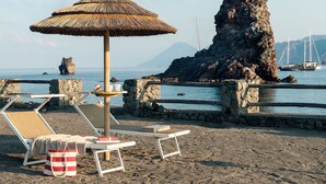 Egy strand, ahol napozóágyak, szalmatetejű napernyő és sziklás partszegély található.