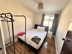 A bedroom with a bed, a bedside table, a window with curtains, and a hanging light fixture. - Large & Cozy 5-bed House in a Quiet Dagenham Area (Dagenham)