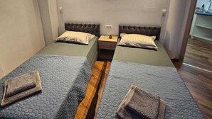 Ein Zimmer mit zwei Betten, Holzboden, Klimaanlage, einem an der Wand hängenden Bild und einem kleinen Tisch mit Lampe.