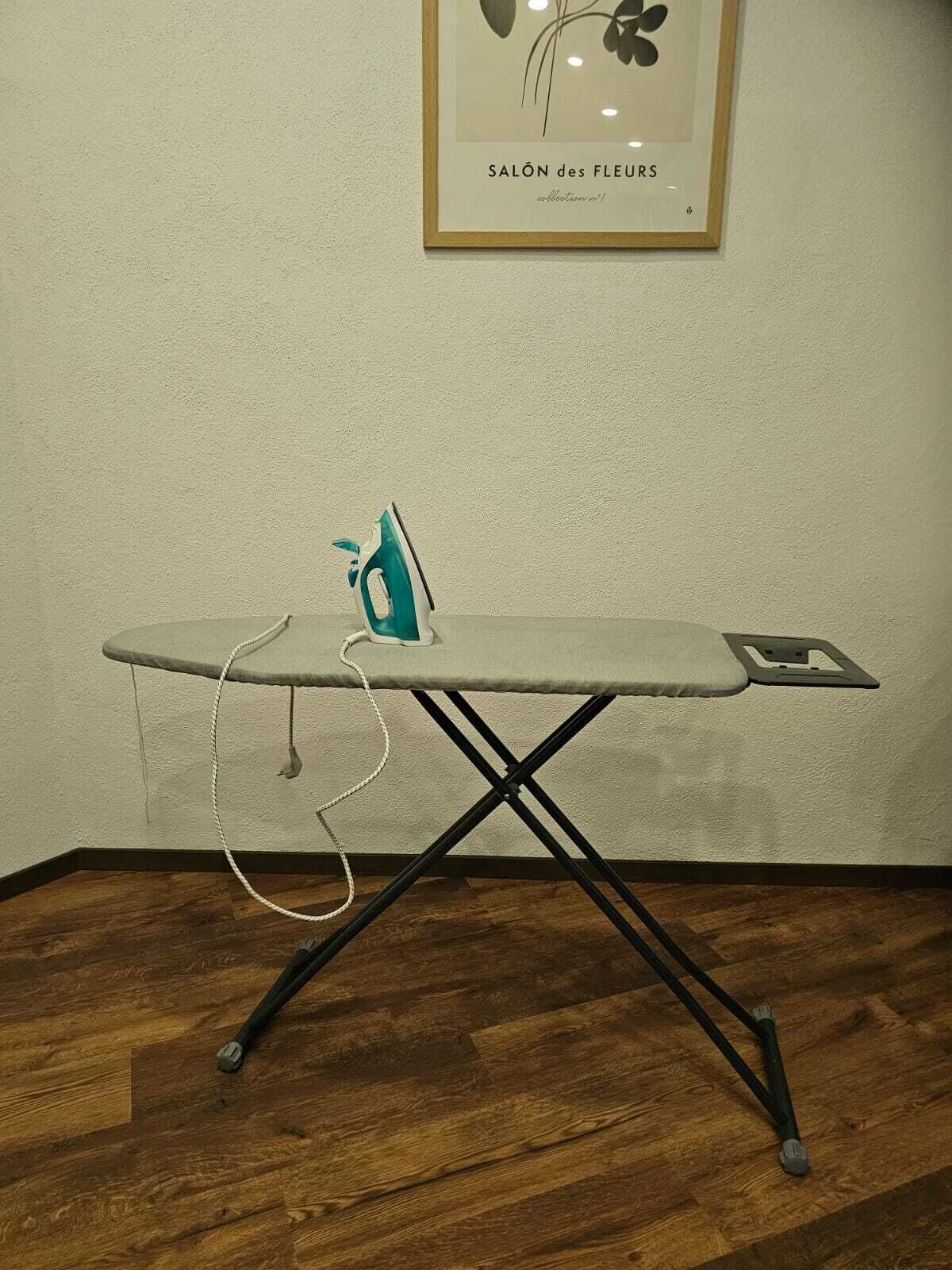 A small, foldable ironing board with an iron and a cord, placed on a wooden floor against a plain wall.