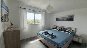 Et soveværelse med en seng, et natbord, et vindue med gardiner, et billede på væggen og en lampe i loftet.