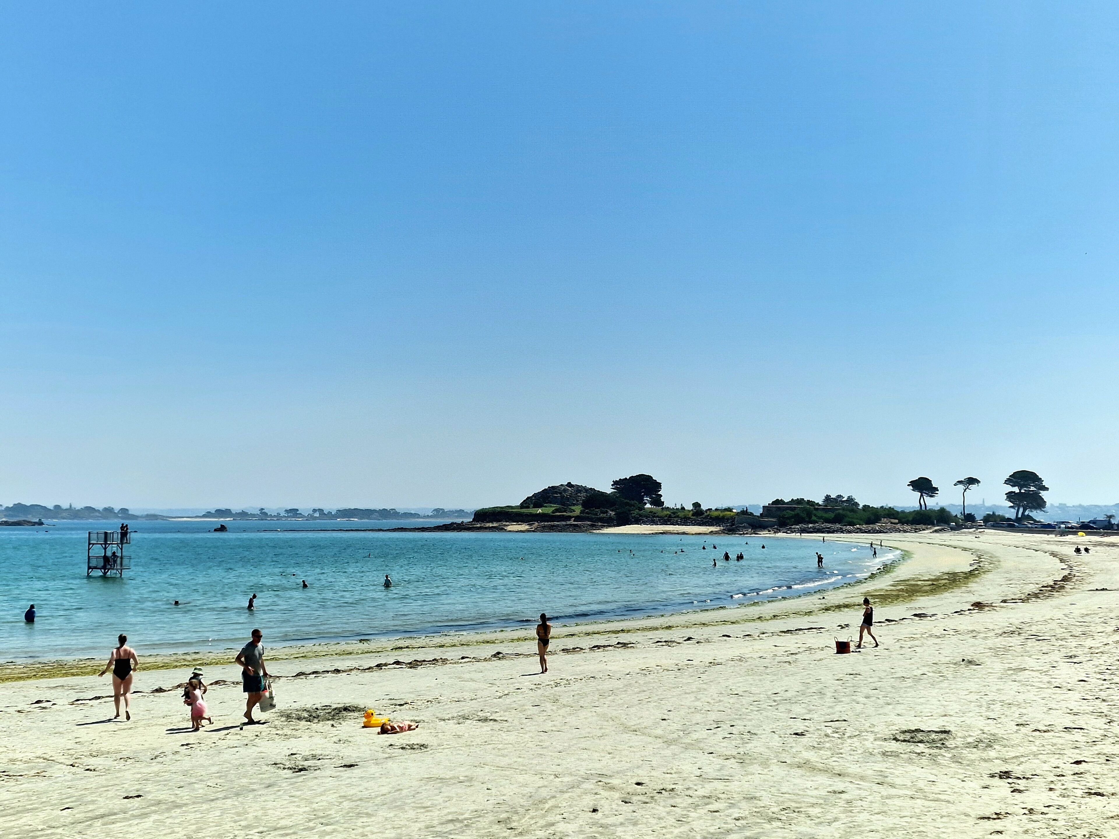A beach with people walking and swimming, a lifeguard tower, and a sandy shore with trees in the background.