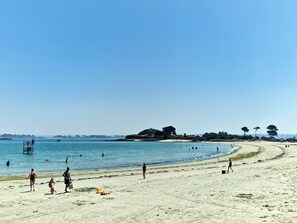 Une plage où l’on voit des gens se promener et nager, une tour de surveillance des sauveteurs, et une rive sablonneuse avec des arbres en arrière-plan.
