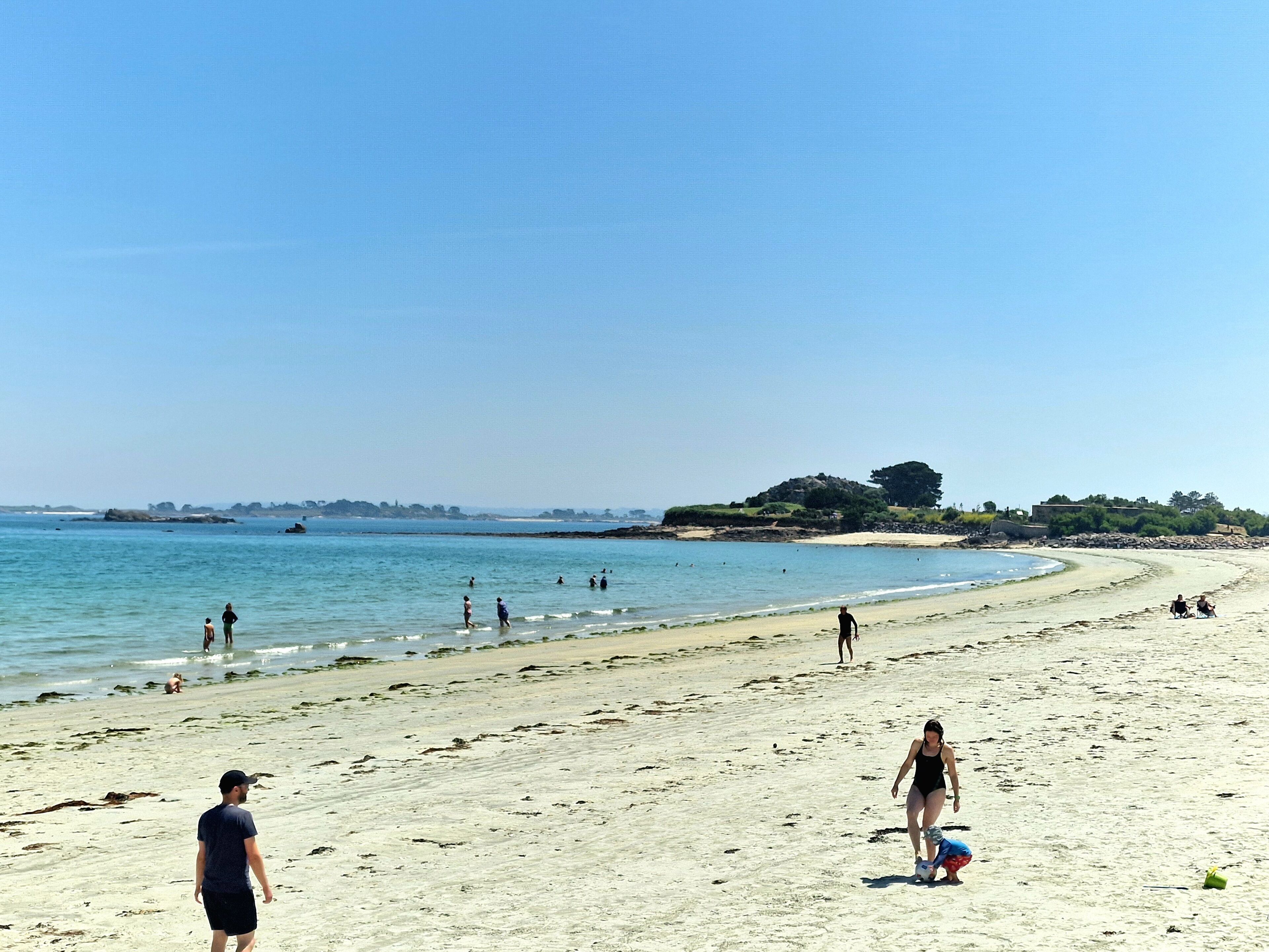 A beach with people walking and playing, a clear sky, and a distant hill with trees.