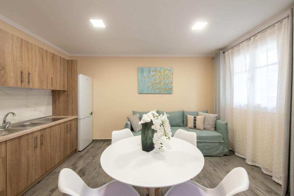 A modern kitchen with wooden cabinets, a white countertop, and a built-in oven. There is a dining area with a white table and chairs, and a sofa with a green cushion. A painting is on the wall above the sofa.