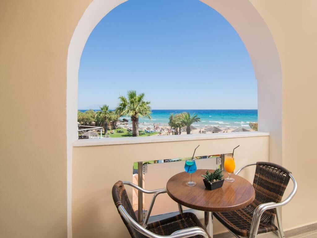 A balcony with a view of the beach, two wicker chairs, a small table with drinks, and a potted plant.