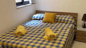 A bed with a checkered bedspread, two rolled towels, a wooden headboard, a nightstand with a lamp, and a window with curtains.