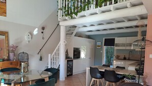 Una cocina moderna con gabinetes blancos, un comedor con mesa y sillas de vidrio, y una escalera con barandilla blanca.
