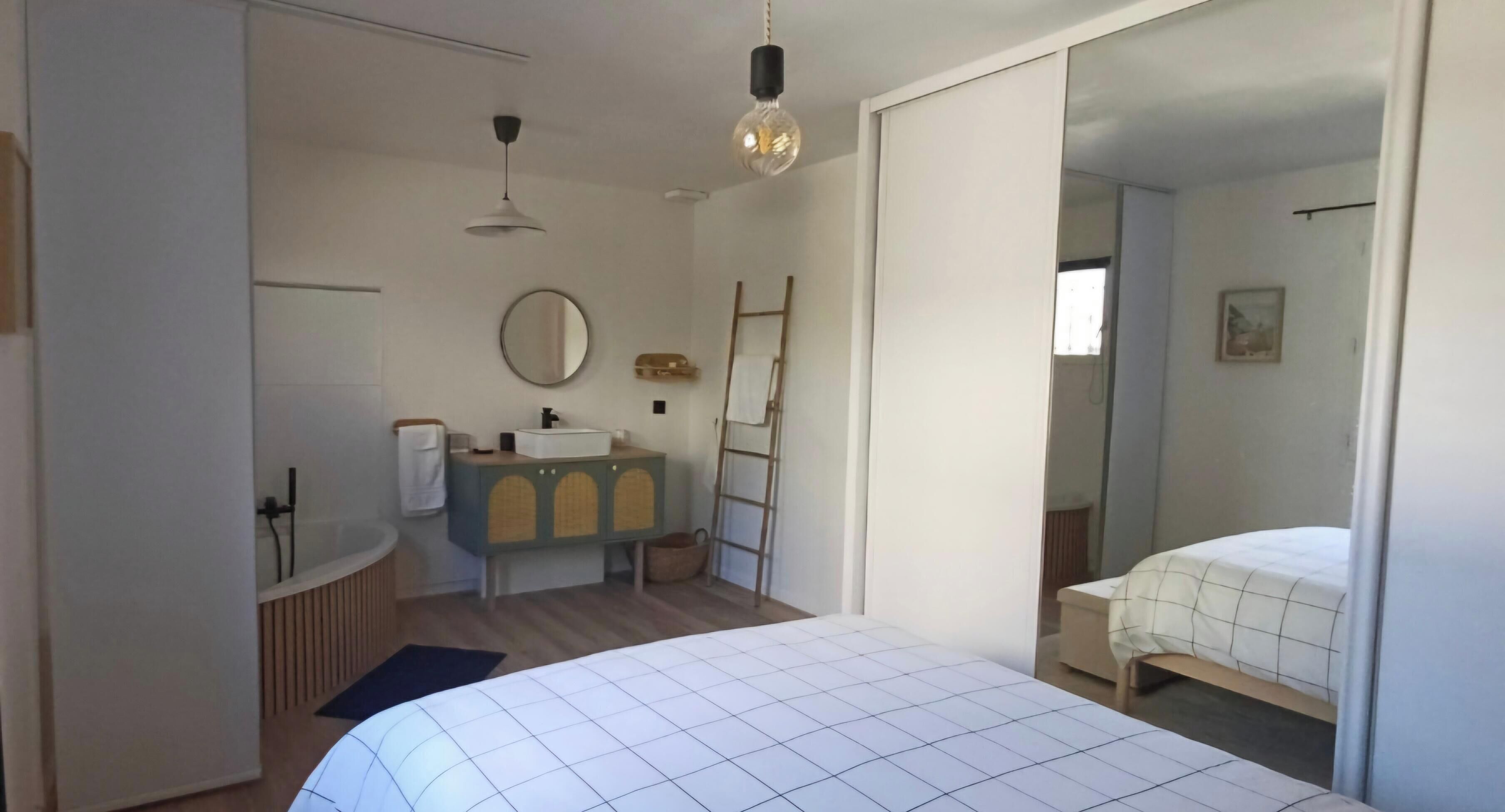 Een slaapkamer met een bed, nachtkastje, spiegel, kast en een badkamer met bad en wastafel.