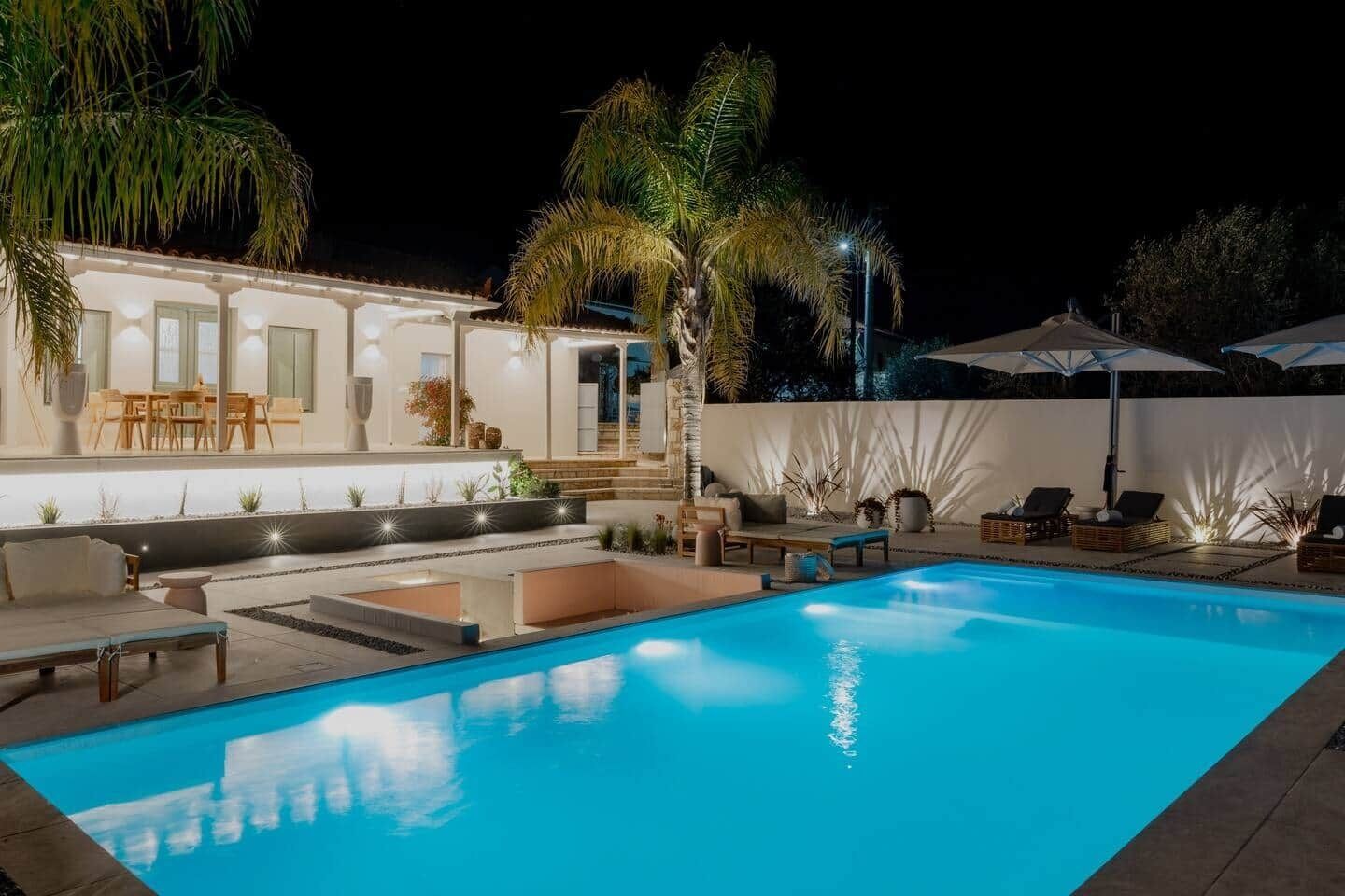 Una piscina con lettini e una zona patio, circondata da palme e un edificio bianco.