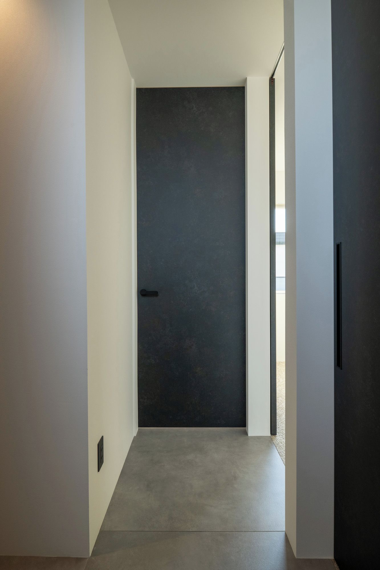 Un pasillo que conduce a una puerta oscura con un picaporte sencillo, un interruptor de luz en la pared y una pequeña ventana a la derecha.