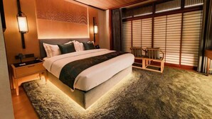 A modern hotel room with a large bed, a nightstand, a wooden headboard, a seating area with a chair, and a large window with blinds. - Tsuruyoshikawa onsen hotel (Manazuru)